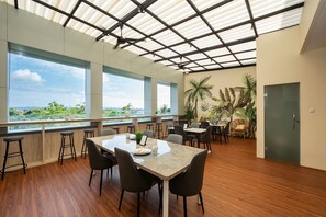 A modern dining area with a large table, multiple chairs, and a view of greenery through large windows. - Cove Tripuri House Bali (Tuban)