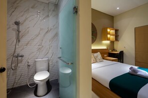 A bathroom with a marble wall, a toilet, and a shower. The room has a bed with a green blanket and a wooden headboard. - Cove Tripuri House Bali (Tuban)