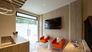A modern living room with a flat-screen TV mounted on the wall, two orange armchairs, a coffee table, and a wooden reception desk. - Cove Tripuri House Bali (Tuban)