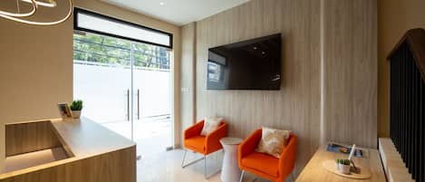 A modern living room with a flat-screen TV mounted on the wall, two orange armchairs, a coffee table, and a wooden reception desk.