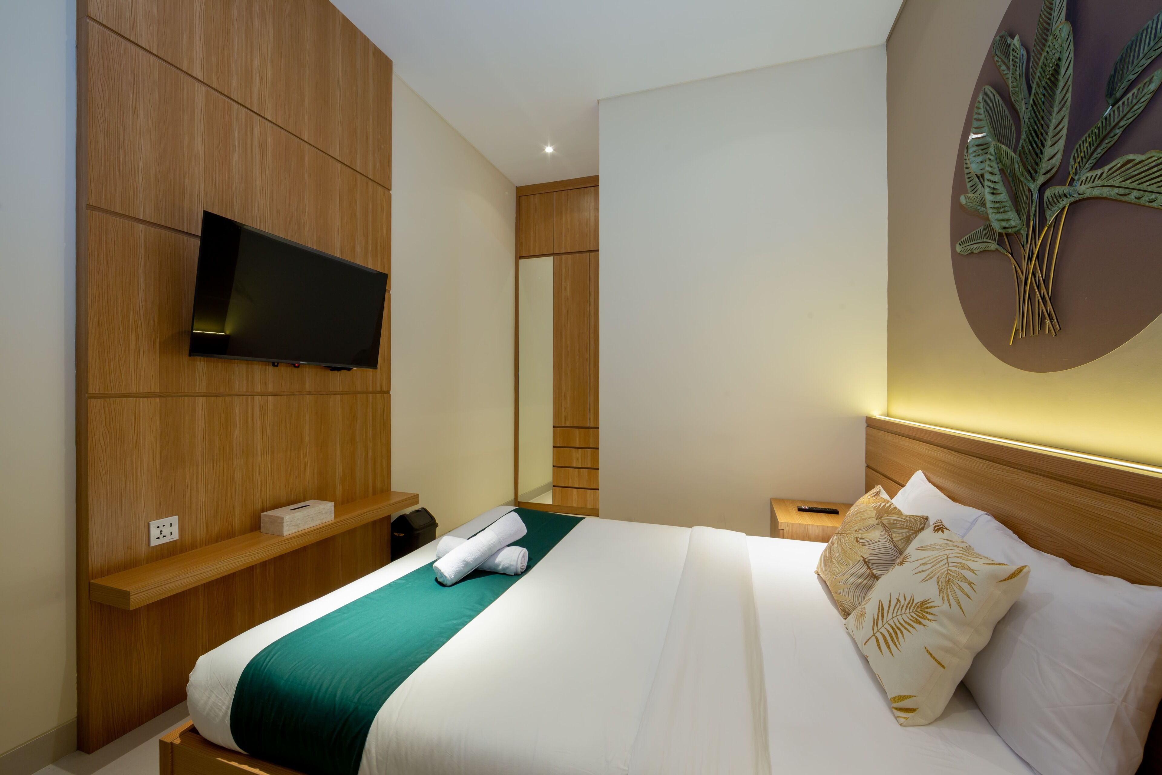Een hotelkamer met een bed, een aan de muur bevestigde televisie, een houten hoofdeinde, een nachtkastje en een wanddecoratie met een plant.