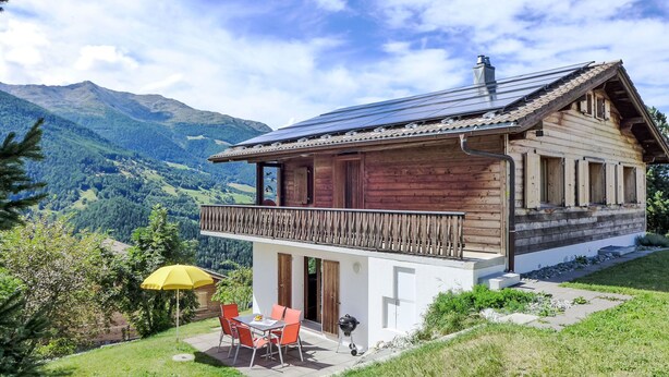 Een houten chalet met balkon, een terras met tafel en stoelen, en uitzicht op bergen.