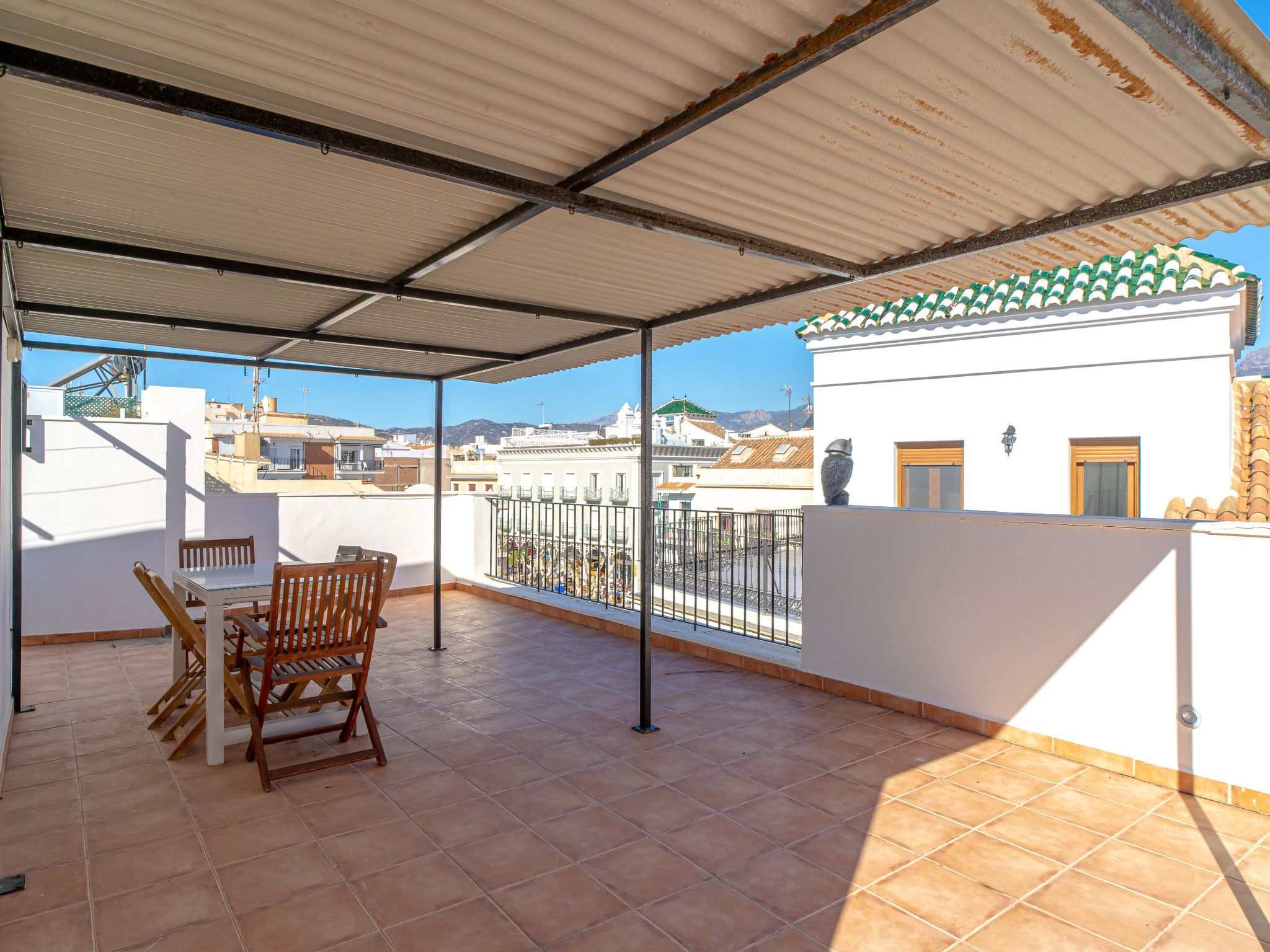 A covered patio with a table and chairs, overlooking a cityscape with buildings and a clear sky.