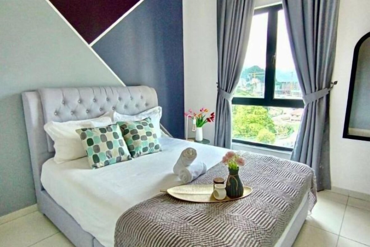 Una cama bien tendida con un cabecero capitonado, una mesita de noche con un florero y una bandeja, y una ventana con cortinas.