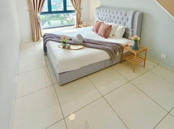 A neatly made bed with a grey headboard, a bedside table with a vase, and a view of the outdoors through a window. - Emerald Sky Suites l The Horizon Ipoh l by Grab A Stay l 1-8pax (Ipoh)