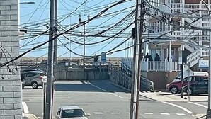 Straßenansicht mit einem weißen Auto am Straßenrand, einem schwarzen Auto im Vordergrund und einem Gebäude mit einem Balkon im Hintergrund.