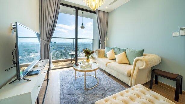 A modern living room with a large window, a white sofa, a round coffee table, and a television on a stand. - HKG-Vinhomes Sky Lake - Luxury Apartment (Hanoi)