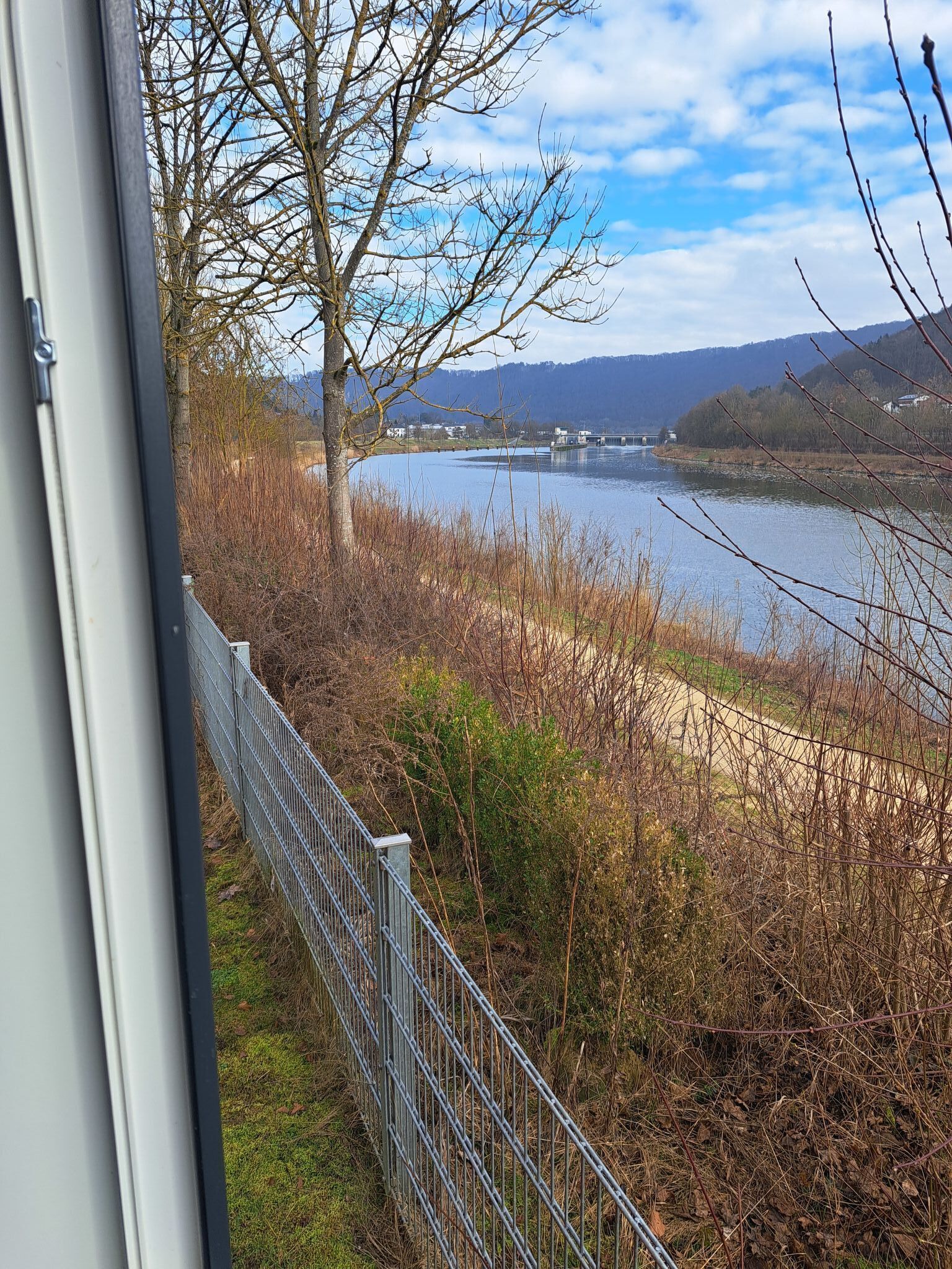Aussicht auf einen Fluss mit einer Brücke, umgeben von Bäumen und einem Metallzaun.