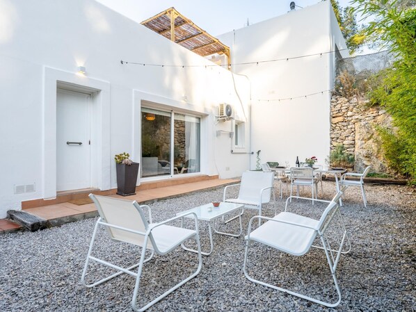 A modern outdoor seating area with white metal chairs and tables, a stone wall, and a white building with a glass door.
