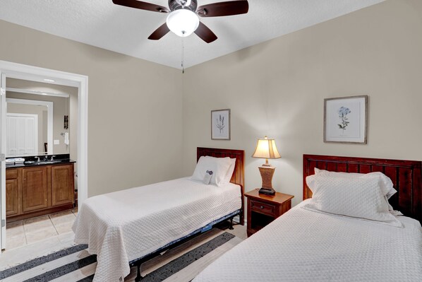A hotel room with two beds, a ceiling fan, a nightstand with a lamp, and framed floral artwork on the walls.