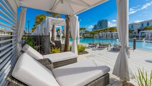 A covered outdoor seating area with white cushions and wicker furniture, overlooking a swimming pool with lounge chairs and umbrellas.