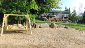 A sandy play area with a wooden swing set, large rocks, and a building in the background. - Rainy Dream Vacation - Spacious accomodations, large yard, incredible view!

 (International Falls)