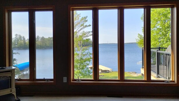 A room with large windows overlooking a lake, a deck with a railing, and a small table with a chair. - Rainy Dream Vacation - Spacious accomodations, large yard, incredible view!

 (International Falls)