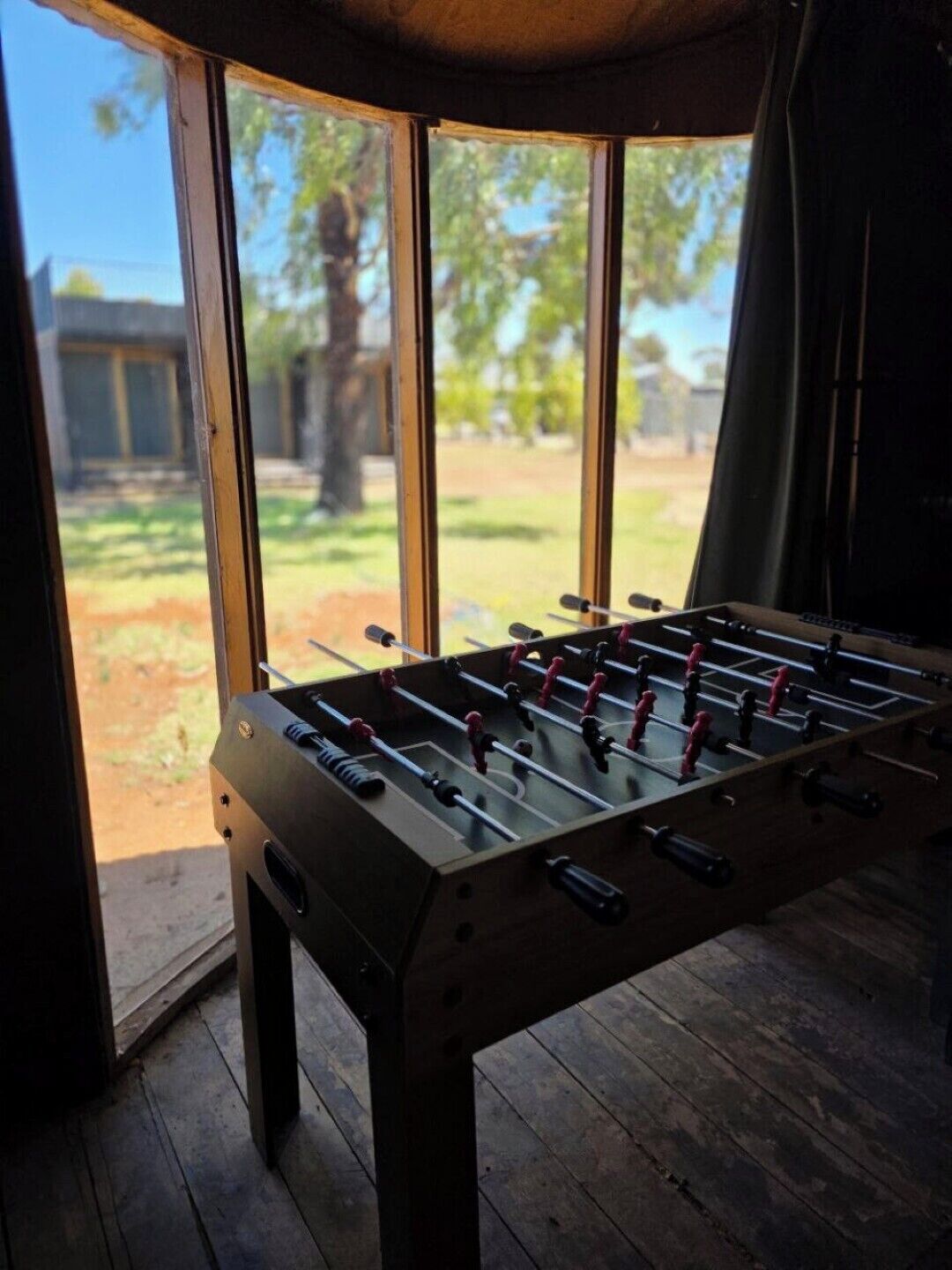 Una mesa de fútbol de mesa en una habitación con ventanas amplias, suelo de madera y vistas a un área verde.