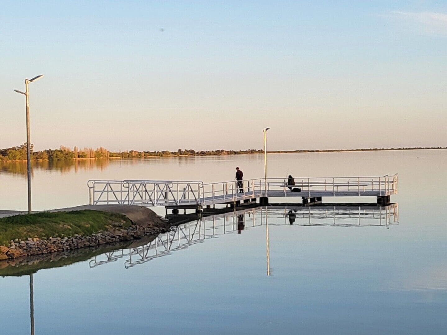 A calm lake with a dock extending into the water, a person standing on the dock, and another person near the water's edge.