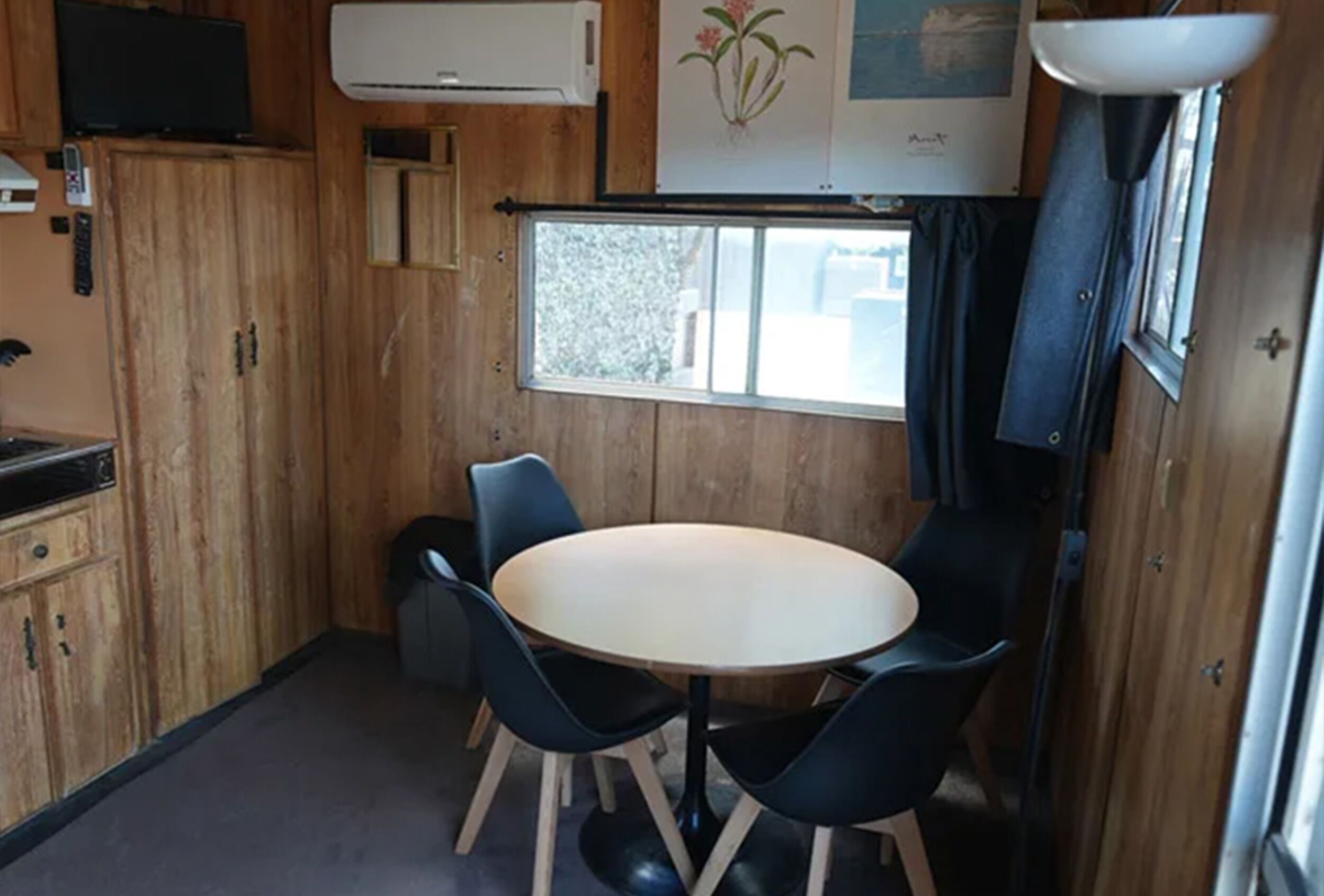 A compact kitchen with a round table and four chairs, wooden cabinets, a small window, and an air conditioning unit.