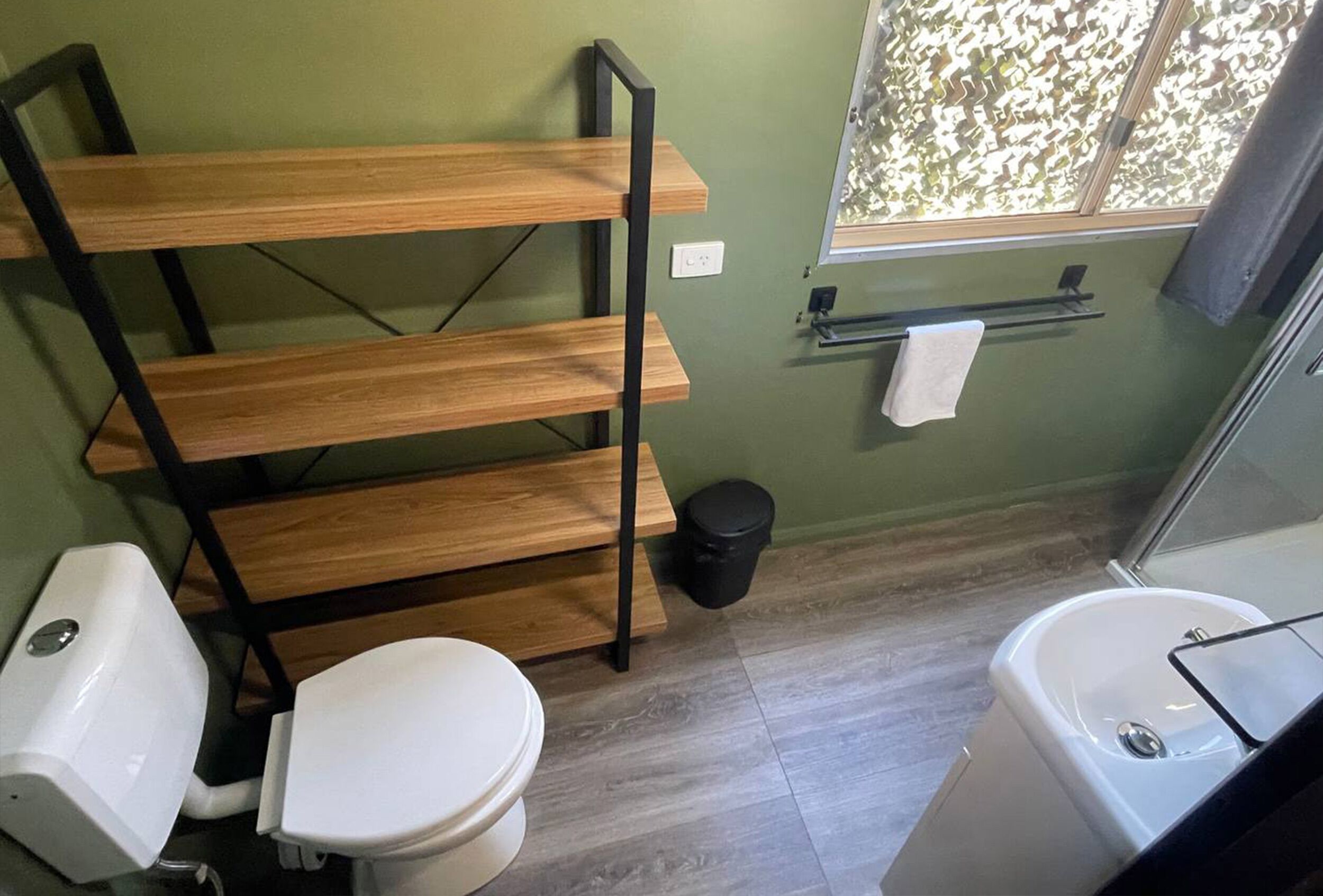A bathroom with a wooden shelf, a toilet, a bidet, a trash bin, and a window with a view.