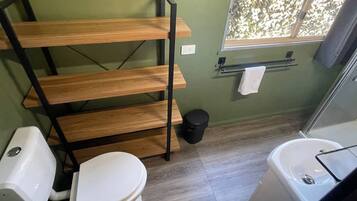 A bathroom with a wooden shelf, a toilet, a bidet, a trash bin, and a window with a view.