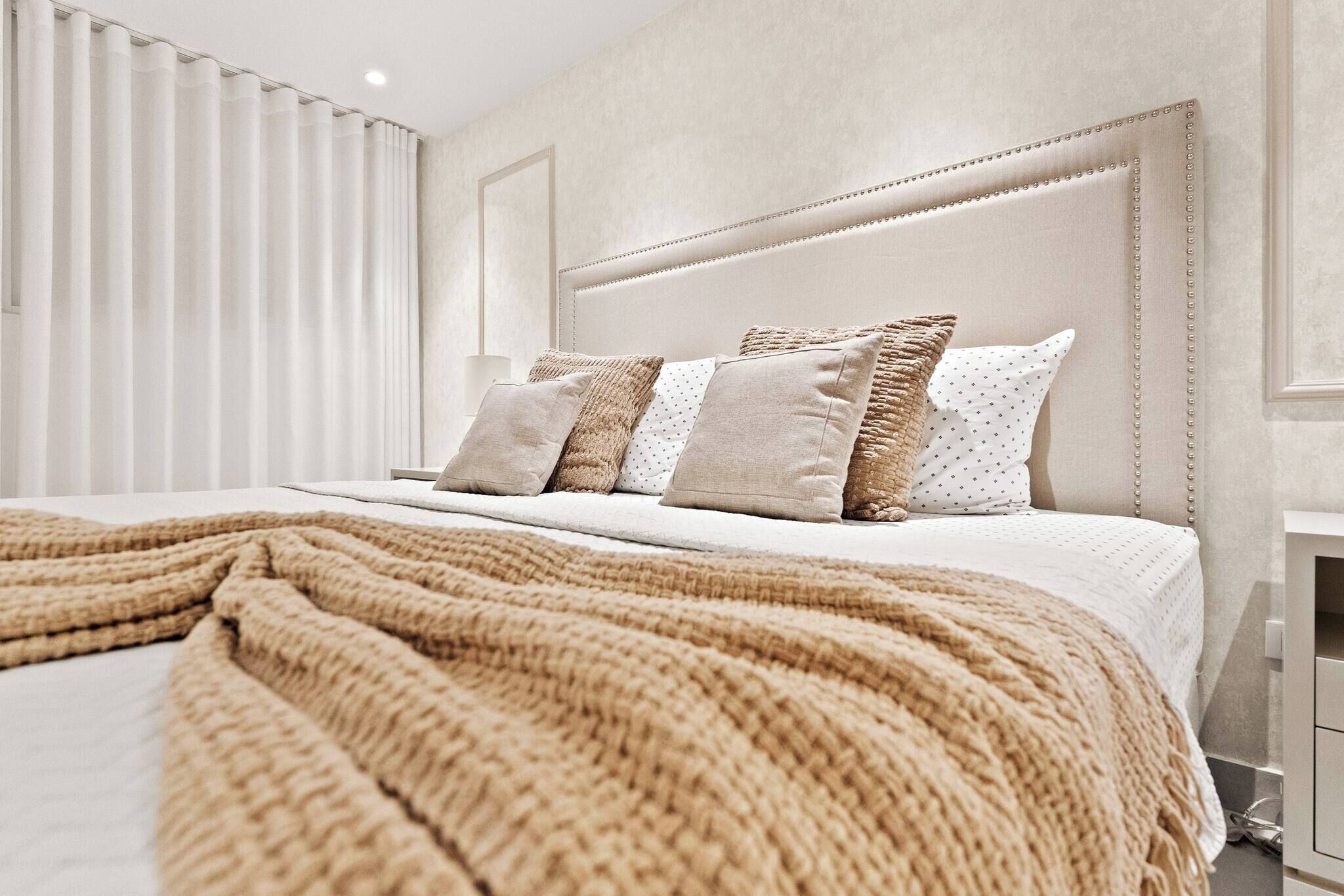 Un letto rifatto con cura, una coperta beige, una testiera con decorazioni e una finestra con persiane verticali.