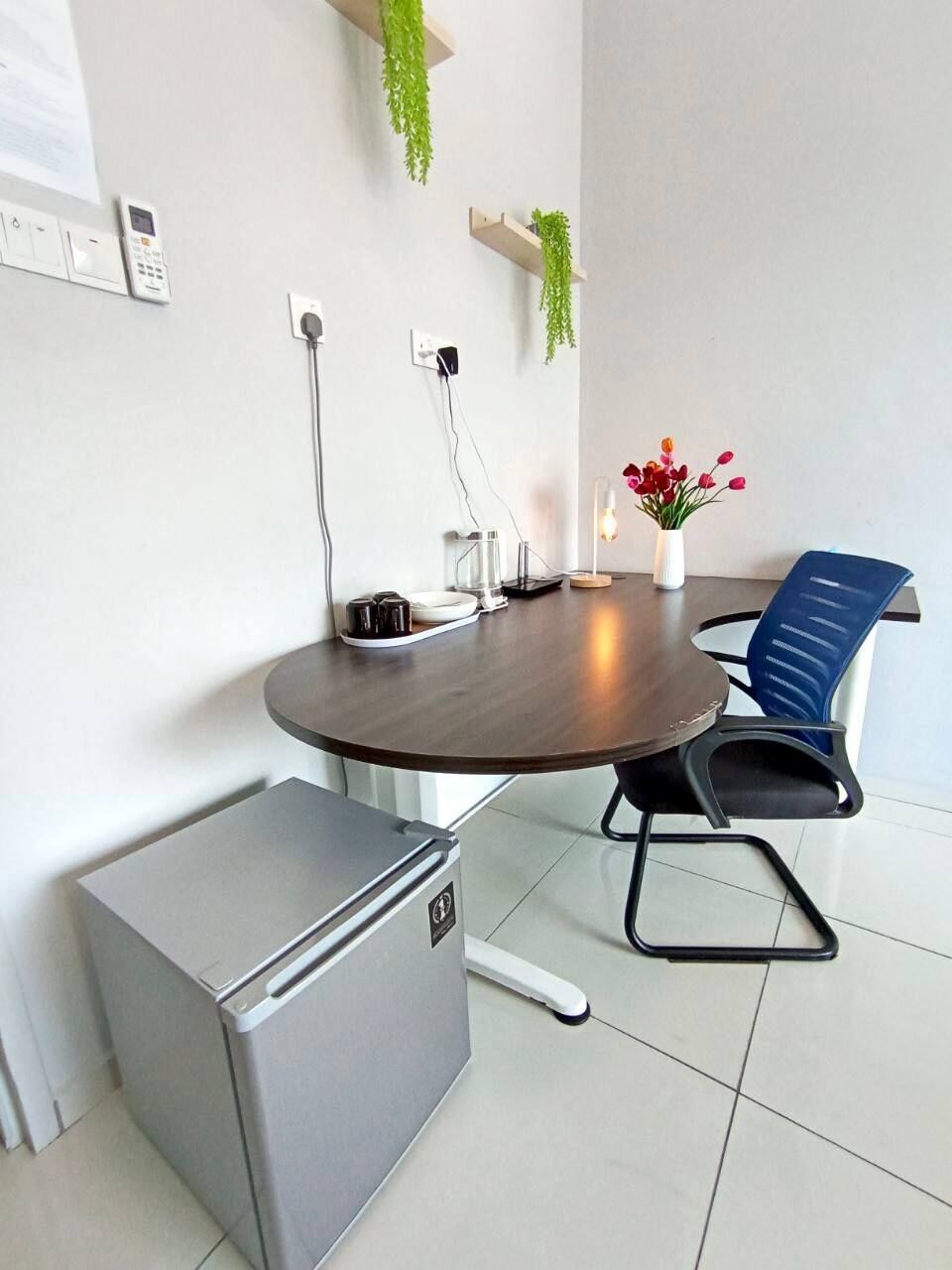 A modern meeting room with a round table, a blue chair, a small refrigerator, a plant, and a vase with flowers.