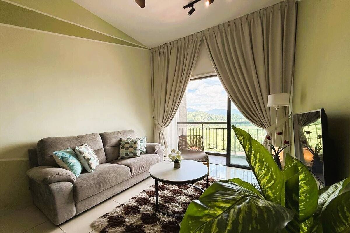 A living room with a sofa, a round coffee table, a large plant, and a balcony with curtains.