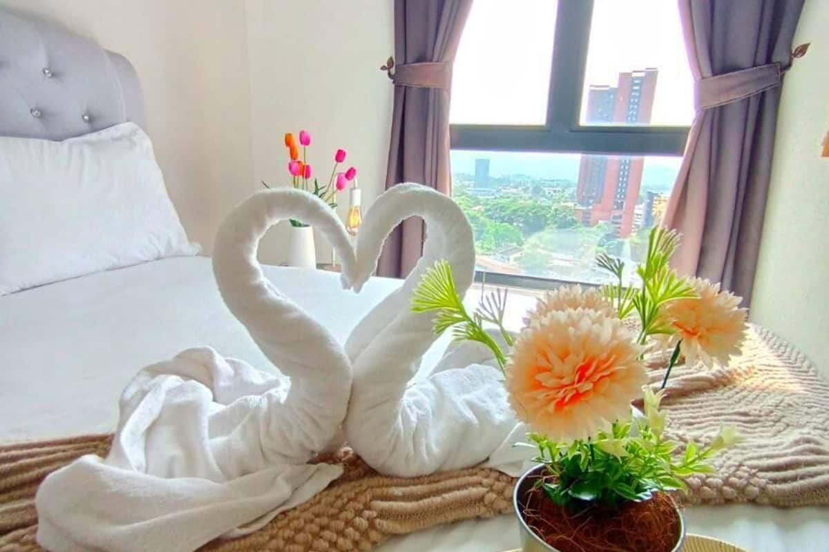 A hotel room with a bed, a window with curtains, a tray with towels, a flower arrangement, and a small potted plant.