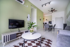 A modern living room with a flat-screen TV, a white round coffee table, a grey sofa, a dining area with a table and chairs, and a kitchen in the background.