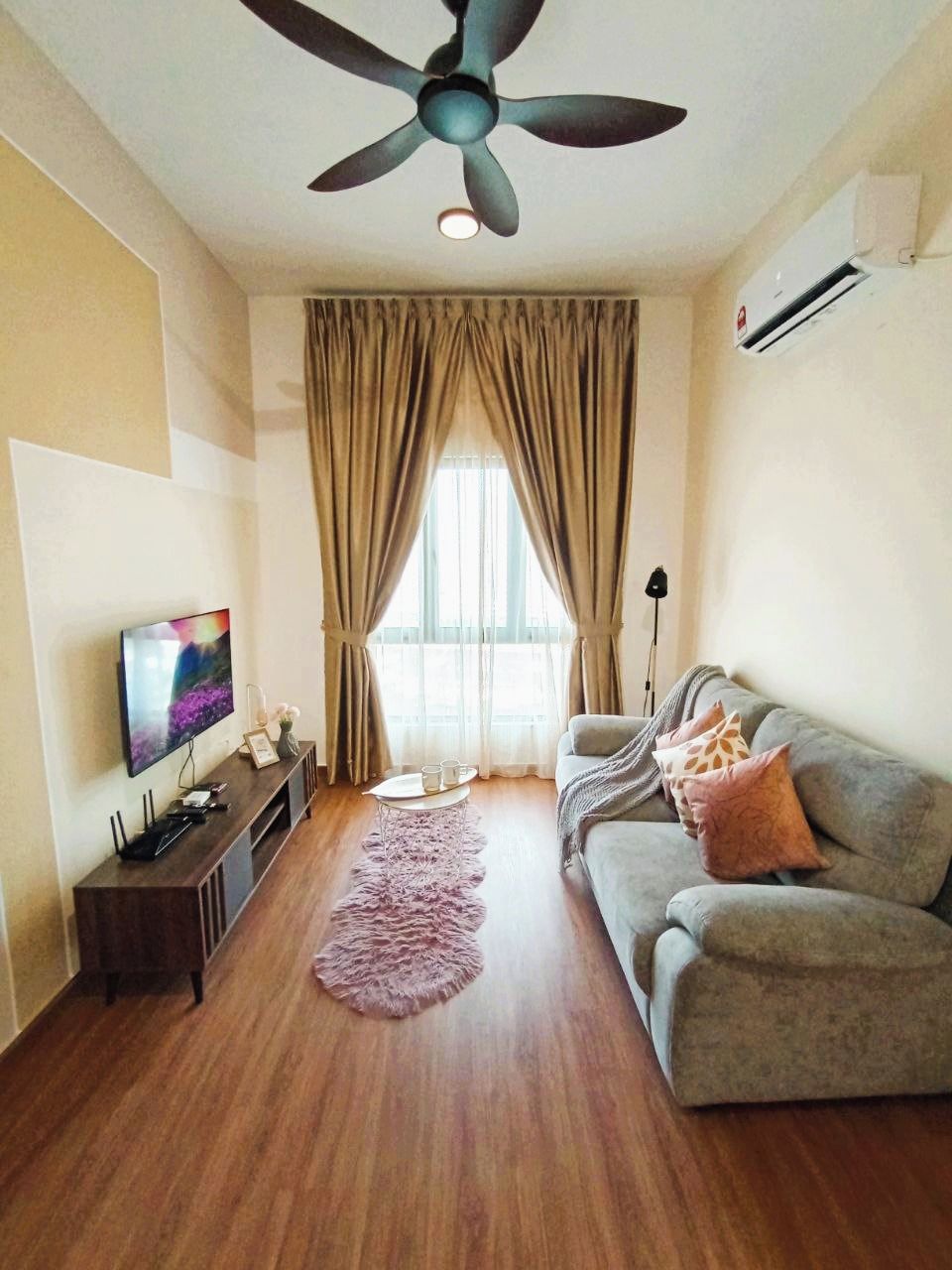 A living room with a ceiling fan, a flat-screen TV, a grey sofa, a wooden floor, and a window with curtains.