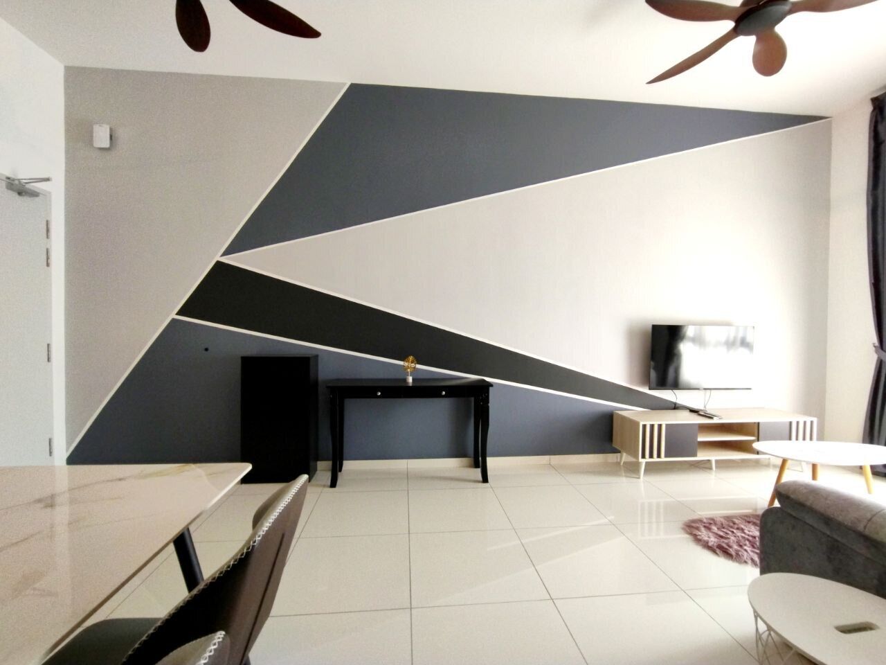 Una sala de estar moderna con un diseño geométrico en la pared, un televisor de pantalla plana, una mesa de centro negra y un sofá gris.
