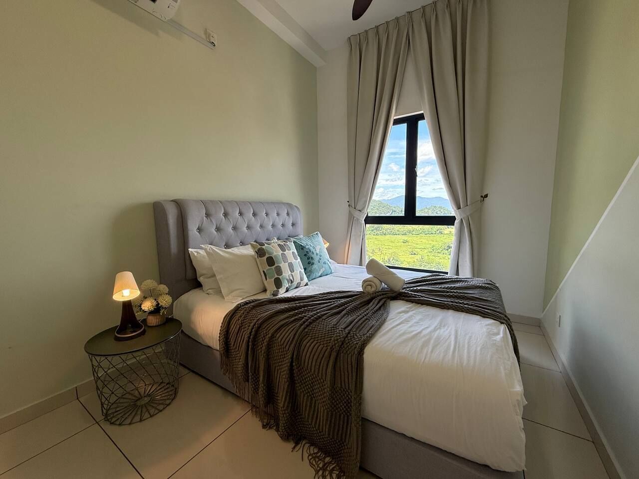 A neatly made bed with a grey headboard, a bedside lamp, and a view of greenery through the window.