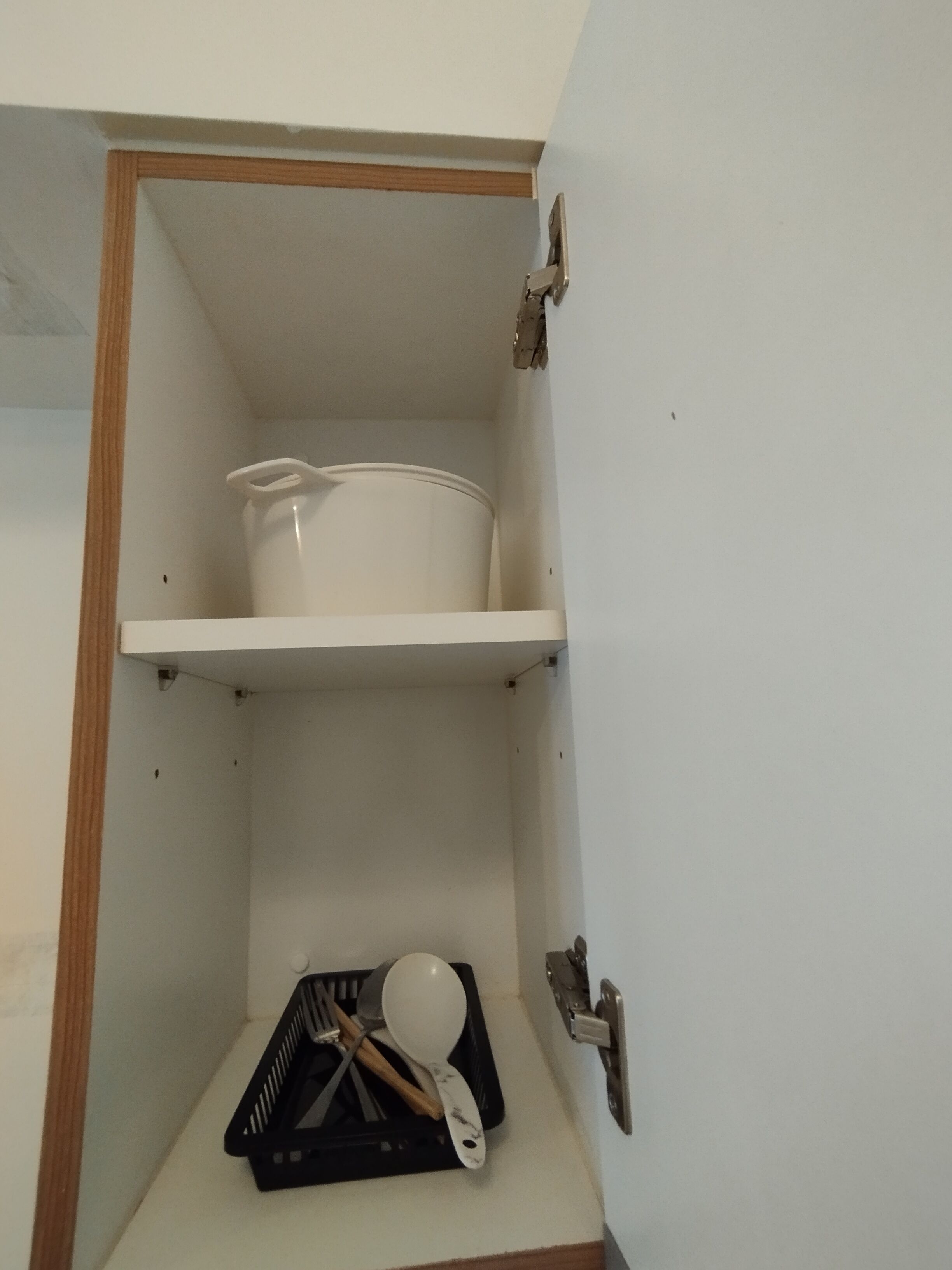 A kitchen cabinet with a white pot on the top shelf and a black tray with utensils on the bottom shelf.