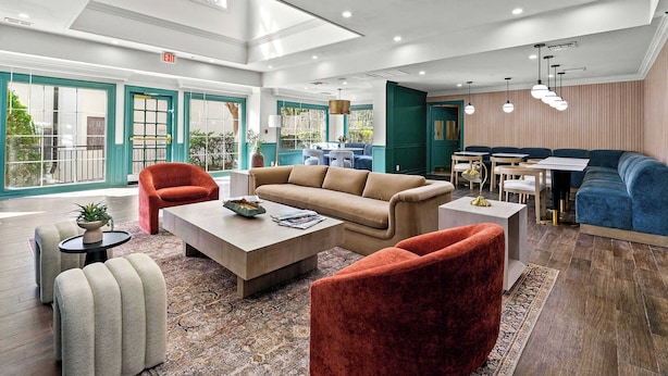 A spacious lobby with a high ceiling, a beige sofa, a red armchair, and a blue sofa. There is a small table with a plant and a magazine.