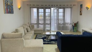 Ein Wohnzimmer mit einem beigefarbenen Ecksofa, einem blauen Sofa und einem Couchtisch. Zwei Fenster mit durchsichtigen Vorhängen, ein Bild an der Wand und ein kleiner Tisch mit Dekoration.