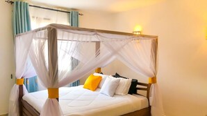 Ein Himmelbett mit weißer Bettwäsche und gelben Akzenten, ein gemustertes Teppich und ein Fenster mit blauen Vorhängen.