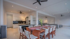 A spacious dining area with a wooden table and white chairs, a kitchen in the background, and ceiling fans. - Olivia Estate (Seminyak)