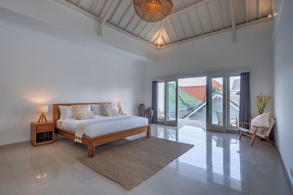 Une chambre spacieuse avec un grand lit, une table de chevet en bois, une lampe sur pied et une vue sur l’extérieur grâce à une porte-fenêtre coulissante.
