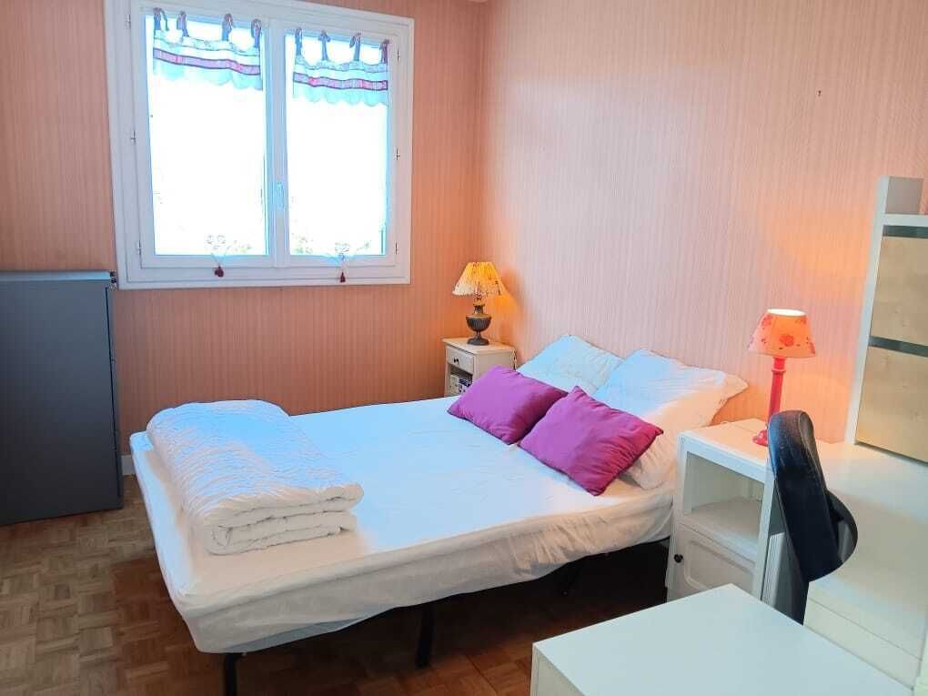 Ein Schlafzimmer mit einem Bett, Kissen, einem Nachttisch, einer Lampe, einem Schreibtisch und einem Fenster mit Vorhängen.