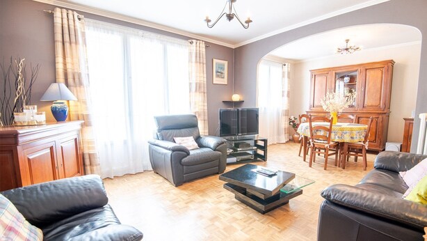 Una sala de estar con sofá, sillón y mesa de centro. Hay un televisor sobre un mueble, una mesa de comedor con sillas y una zona de cocina con armarios.