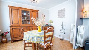 Un comedor con un aparador de madera, una mesa redonda con mantel estampado, sillas, un florero con flores, un radiador y una lámpara fijada a la pared.