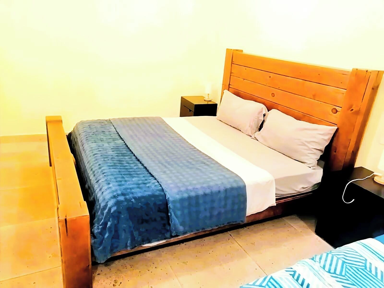 A bed with a wooden headboard, a blue bedspread, and two pillows. There is a nightstand with a lamp beside the bed.