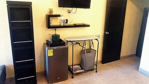 Una stanza compatta con TV a schermo piatto fissata al muro, un piccolo frigorifero, un carrello metallico con un cestino per la spazzatura e una porta.