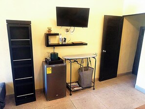 Ett litet rum med en platt-TV som är monterad på väggen, ett litet kylskåp, en metallvagn med en papperskorg och en dörr.