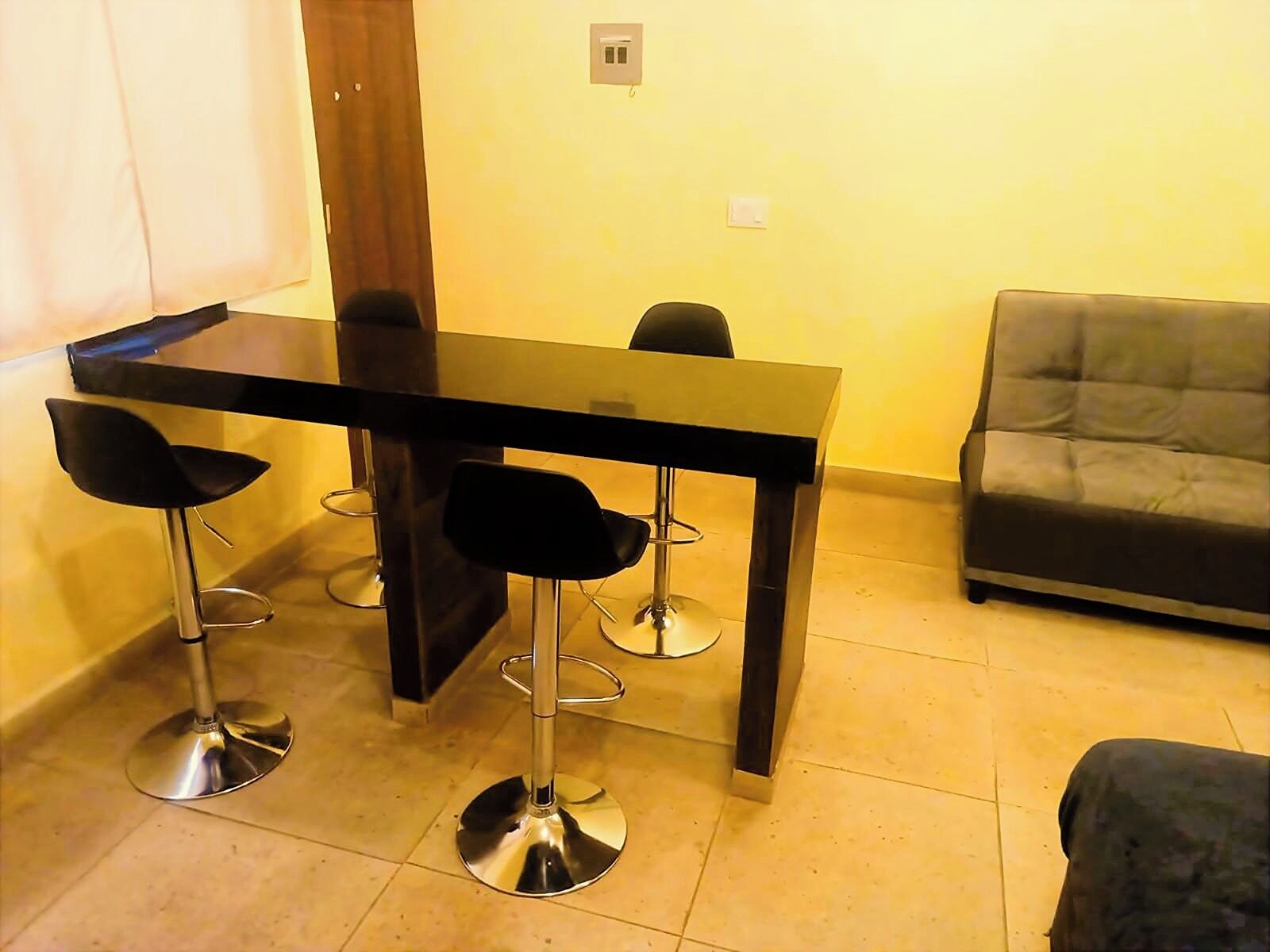 Un coin repas avec une table noire et quatre chaises noires, un canapé gris et un sol carrelé.