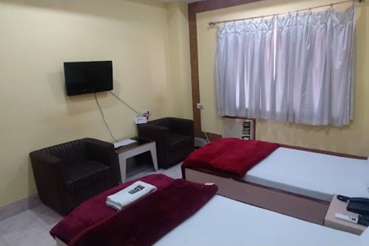 A hotel room with two single beds, a TV mounted on the wall, a small table, a chair, and a window with curtains.