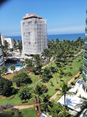 A high-rise building with a pool and palm trees, surrounded by greenery and a view of the ocean. - M3 Riviera Inn Residence 84 (Bertioga)
