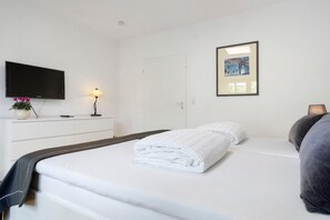 Uma cama bem arrumada com roupa de cama branca, uma TV de tela plana na parede, um abajur e um quadro na parede.