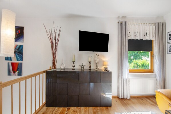 A modern living room with a dark wooden console, a mounted TV, a window with curtains, and artwork on the walls.