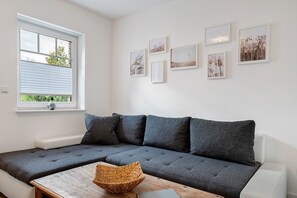 A modern living room with a grey sectional sofa, a wooden coffee table, and a window with blinds. - Holiday Home 'Melody' with Private Terrace and Wi-Fi (Ralswiek)