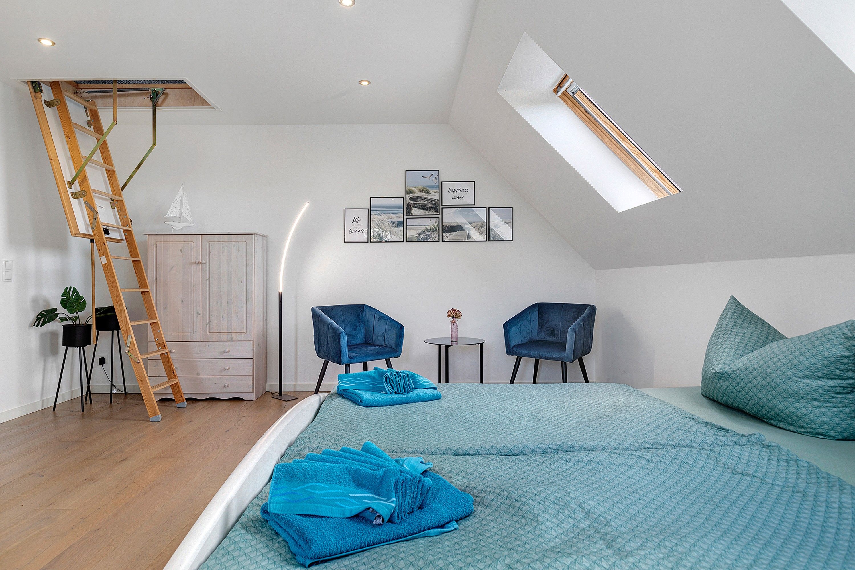 Ein Schlafzimmer mit Schräge, einem Schlafplatz in der Höhe, einer Leiter, zwei Stühlen, einem kleinen Tisch und Wandbildern.