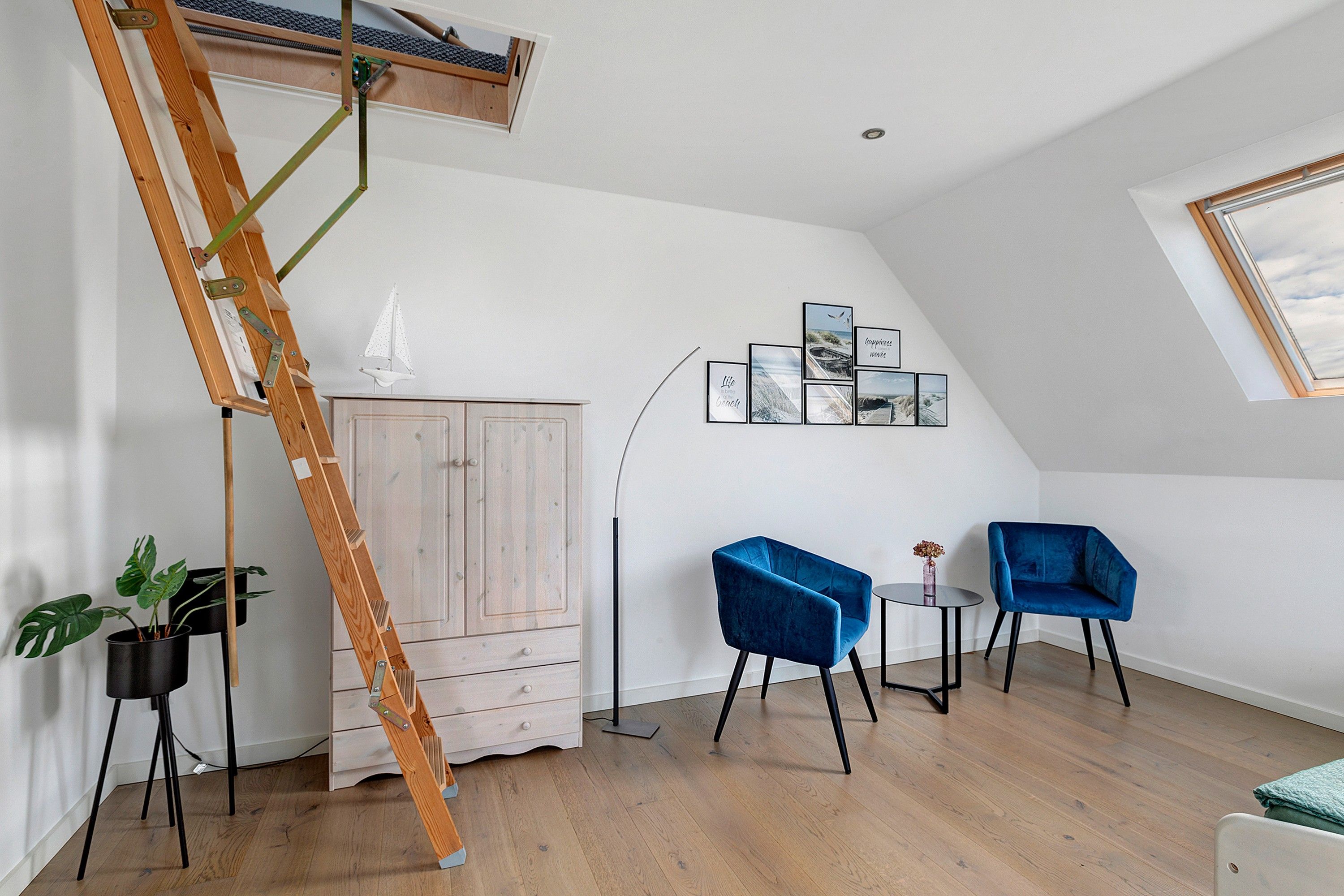 A room with a loft bed, a wooden ladder, a white cabinet, two blue chairs, a small table with a vase, and a wall with framed pictures.